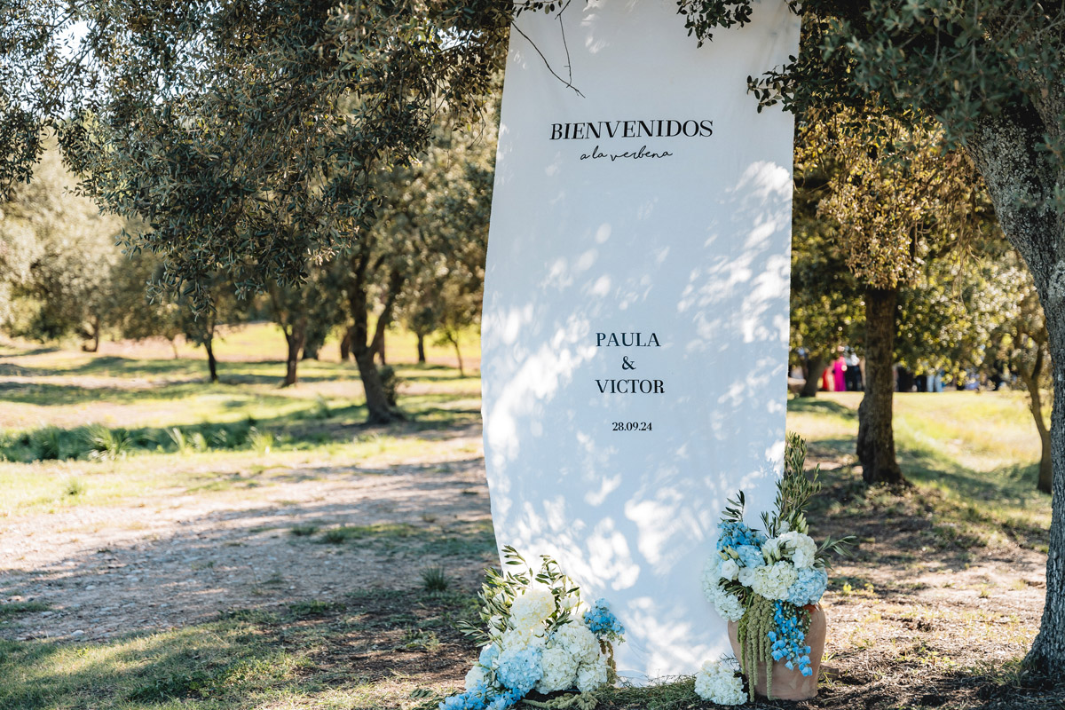 Cartel de bienvenida en tela colgada de un árbol, decorado con hortensias y delphinium en tonos blancos y azules